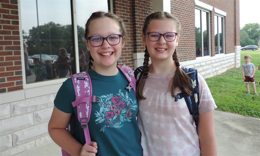 Two girls smile as they stand in front of their elementary school